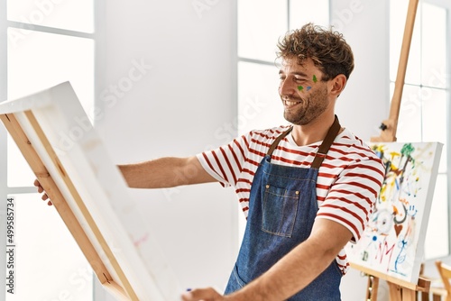 Young hispanic man smiling confident holding canvas at art studio