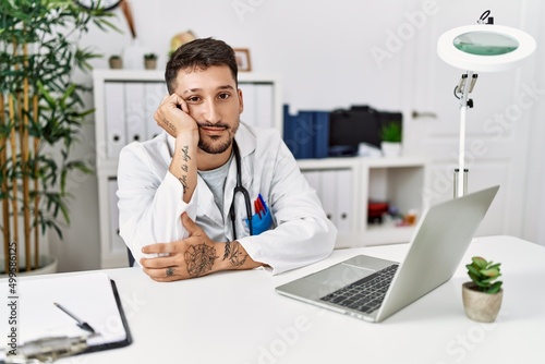 Young doctor working at the clinic using computer laptop thinking looking tired and bored with depression problems with crossed arms.