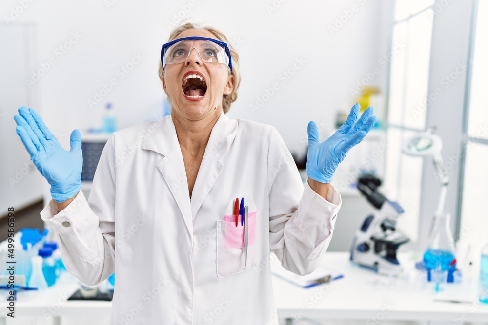 Middle age blonde woman working at scientist laboratory crazy and mad shouting and yelling with aggressive expression and arms raised. frustration concept. Middle age blonde woman working at scientist laboratory crazy and mad shouting and yelling with aggressive expression and arms raised. frustration concept.