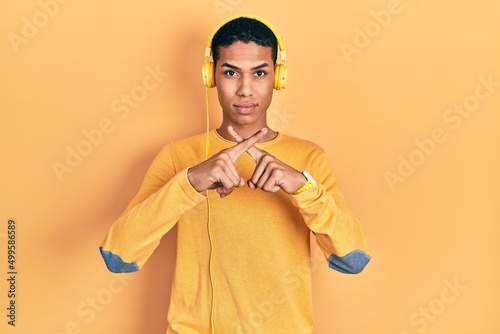 Young african american guy listening to music using headphones rejection expression crossing fingers doing negative sign