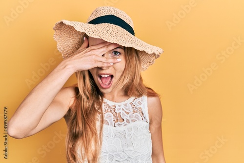 Young caucasian woman wearing summer hat peeking in shock covering face and eyes with hand, looking through fingers with embarrassed expression.