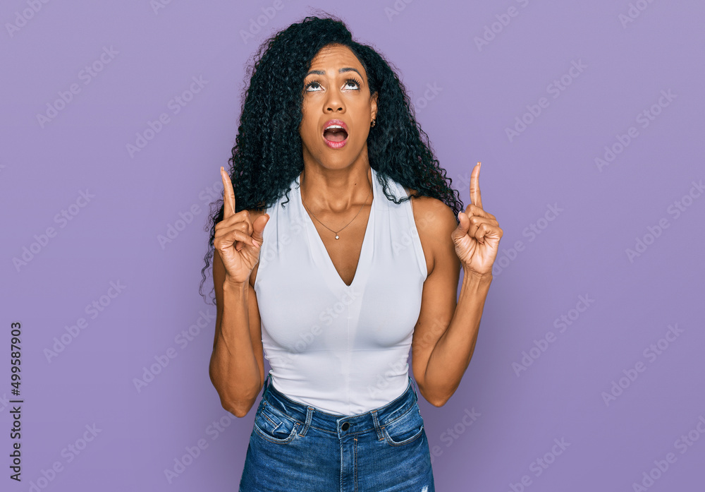 Middle age african american woman wearing casual style with sleeveless shirt amazed and surprised looking up and pointing with fingers and raised arms. Middle age african american woman wearing casual style with sleeveless shirt amazed and surprised looking up and pointing with fingers and raised arms.