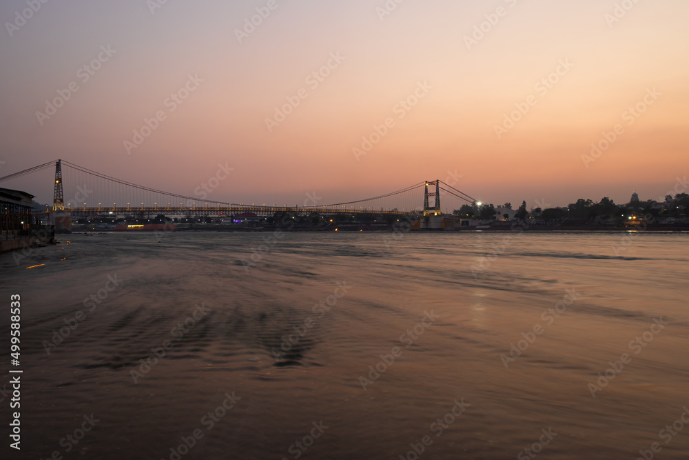 iron suspension bridge with sunset dramatic sky and blurred flowing river water at evening