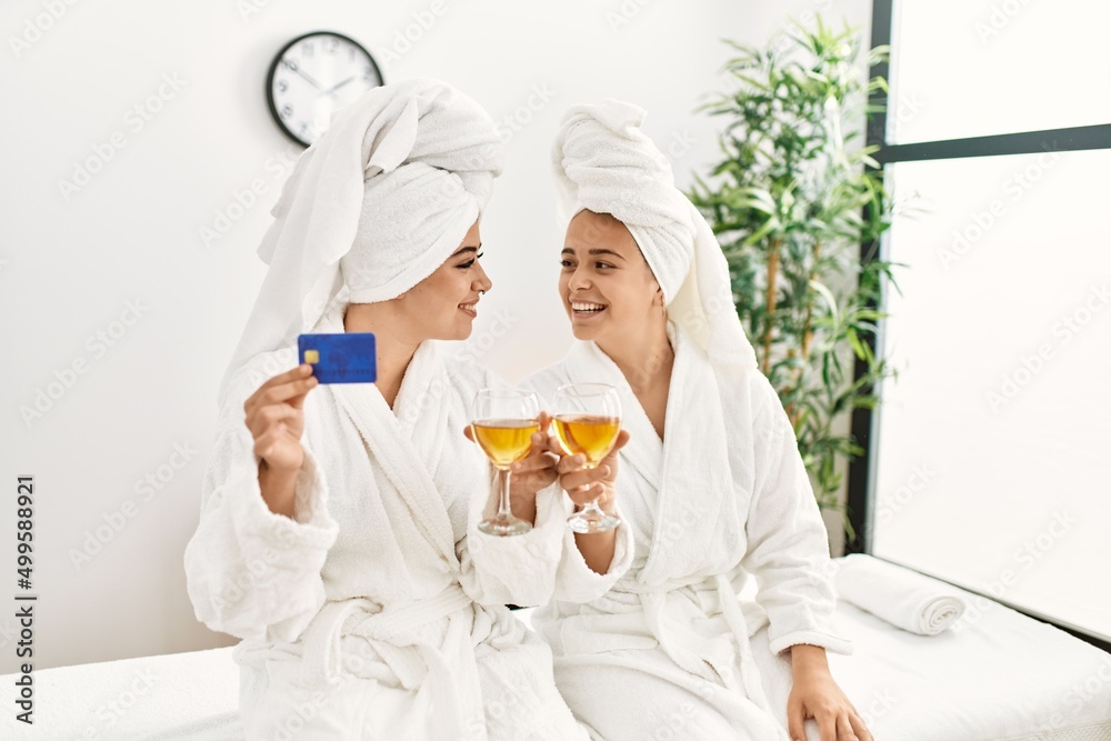 Woman couple holding credit card and toasting with champagne sitting on massage board at beauty center Woman couple holding credit card and toasting with champagne sitting on massage board at beauty center