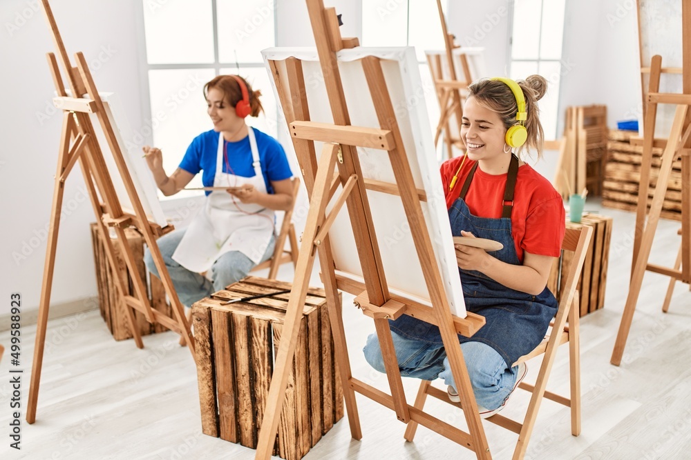 Two artist student women smiling happy and listening to music painting at art school.