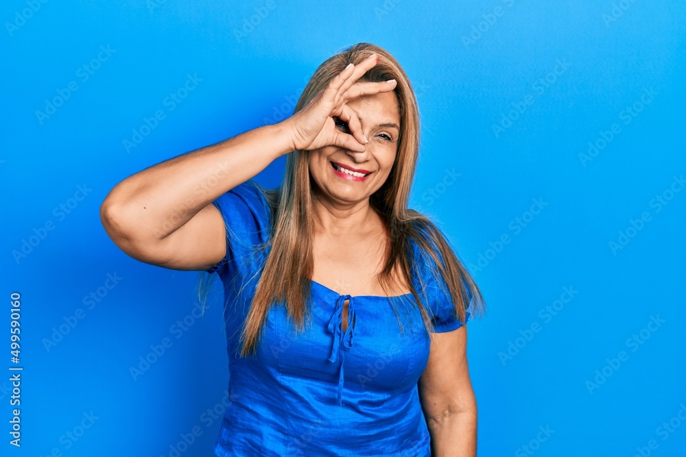 Middle age hispanic woman wearing casual clothes smiling happy doing ok sign with hand on eye looking through fingers