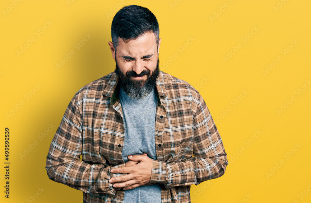 Hispanic man with beard wearing casual shirt with hand on stomach because nausea, painful disease feeling unwell. ache concept. Hispanic man with beard wearing casual shirt with hand on stomach because nausea, painful disease feeling unwell. ache concept.