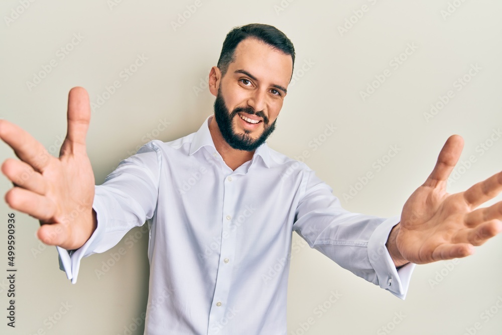Young man with beard wearing business white shirt looking at the camera smiling with open arms for hug. cheerful expression embracing happiness. Young man with beard wearing business white shirt looking at the camera smiling with open arms for hug. cheerful expression embracing happiness.