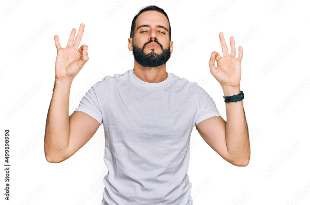 Young man with beard wearing casual white t shirt relaxed and smiling with eyes closed doing meditation gesture with fingers. yoga concept. Young man with beard wearing casual white t shirt relaxed and smiling with eyes closed doing meditation gesture with fingers. yoga concept.