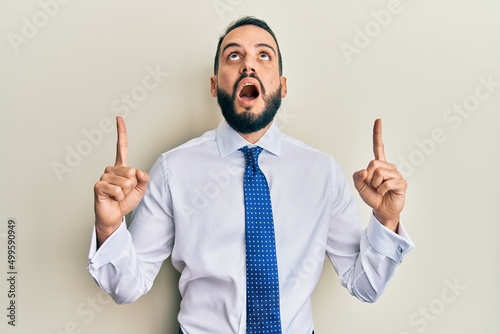 Young man with beard wearing business tie amazed and surprised looking up and pointing with fingers and raised arms.