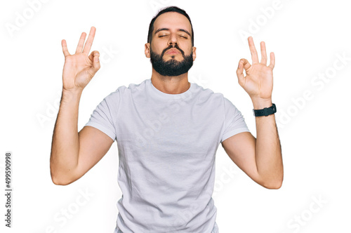 Young man with beard wearing casual white t shirt relaxed and smiling with eyes closed doing meditation gesture with fingers. yoga concept.
