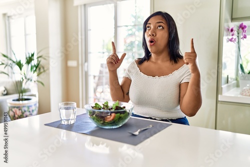 Young hispanic woman eating healthy salad at home amazed and surprised looking up and pointing with fingers and raised arms.
