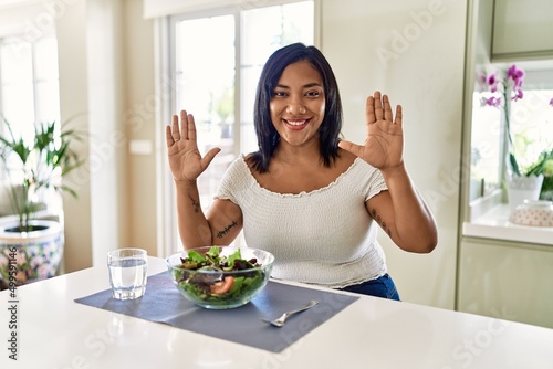 Young hispanic woman eating healthy salad at home showing and pointing up with fingers number ten while smiling confident and happy.