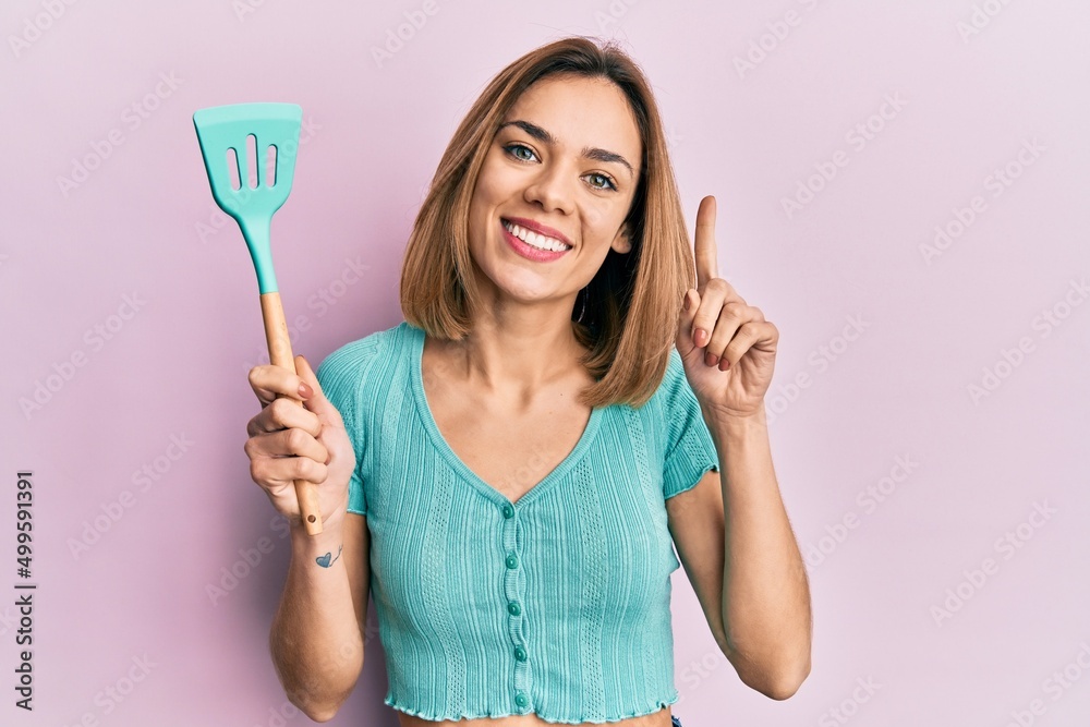 Young caucasian blonde woman holding silicone spatula smiling with an idea or question pointing finger with happy face, number one Young caucasian blonde woman holding silicone spatula smiling with an idea or question pointing finger with happy face, number one