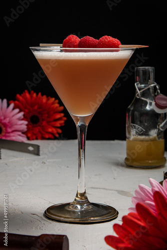 A French Martini cocktail garnished with fresh raspberries on a white background and styled with bar equipment and flowers.