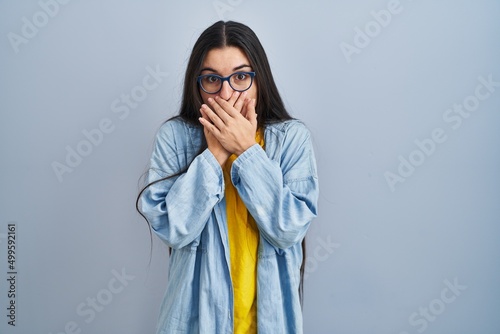 Young hispanic woman standing over blue background shocked covering mouth with hands for mistake. secret concept.