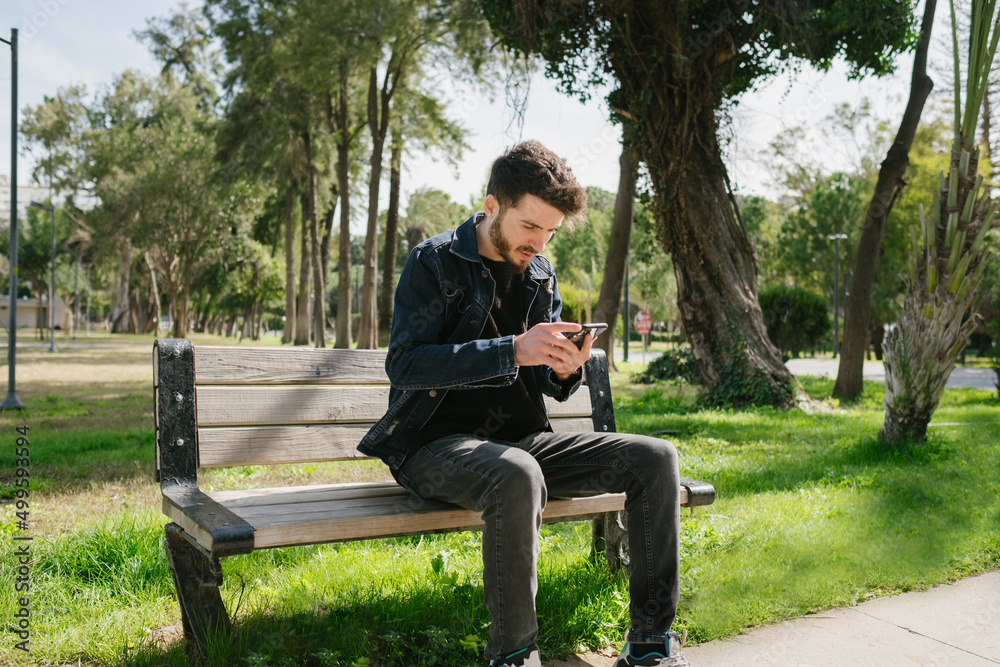 Young man looking at phone at park bench, man in denim jacket looking at black screen, online shopping and internet surfing Young man looking at phone at park bench, man in denim jacket looking at black screen, online shopping and internet surfing