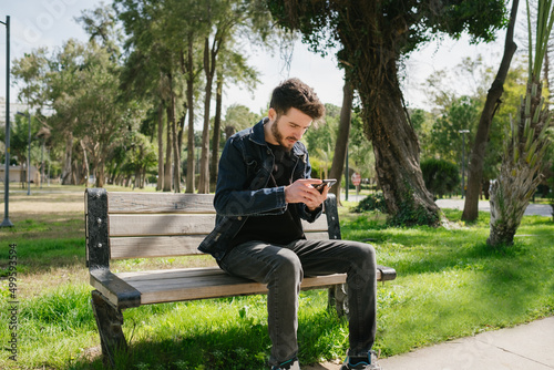 Young man looking at phone at park bench, man in denim jacket looking at black screen, online shopping and internet surfing