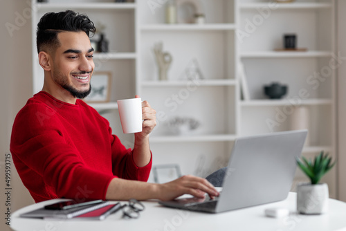 Cheerful attractive young arab businessman with beard in red clothes drinks coffee and types on laptop at workplace