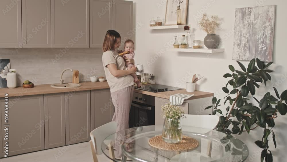 Medium long of young Biracial woman in loungewear holding adorable infant, standing in kitchen at daytime, drinking coffee from mug Medium long of young Biracial woman in loungewear holding adorable infant, standing in kitchen at daytime, drinking coffee from mug