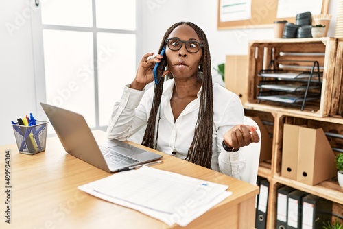 Black woman with braids working at the office speaking on the phone making fish face with lips, crazy and comical gesture. funny expression.