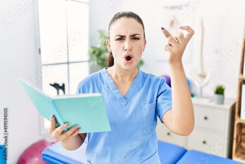 Young physiotherapist woman reading a book at pain recovery clinic annoyed and frustrated shouting with anger, yelling crazy with anger and hand raised