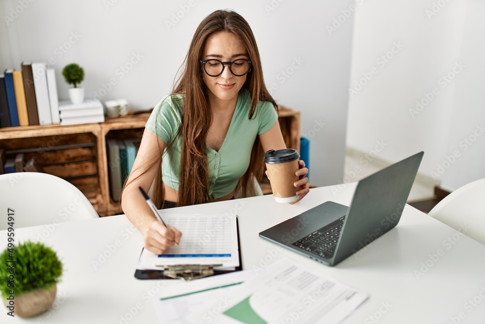 Young hispanic woman using laptop and writing on clipboard sitting on table at home Young hispanic woman using laptop and writing on clipboard sitting on table at home