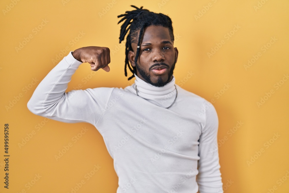 African man with dreadlocks wearing turtleneck sweater over yellow background strong person showing arm muscle, confident and proud of power African man with dreadlocks wearing turtleneck sweater over yellow background strong person showing arm muscle, confident and proud of power