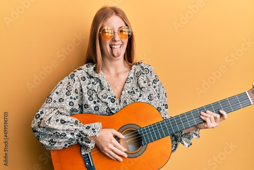 Young irish woman wearing boho style playing classical guitar sticking tongue out happy with funny expression.