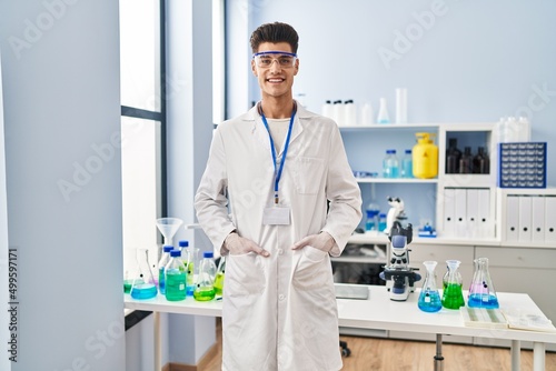 Young hispanic man wearing scientist uniform standing at laboratory