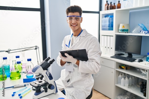 Young hispanic man wearing scientist uniform writing on clipboard at laboratory