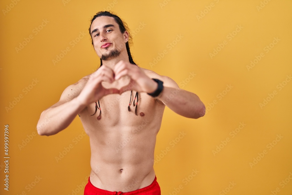 Hispanic man with long hair standing shirtless over yellow background smiling in love doing heart symbol shape with hands. romantic concept.