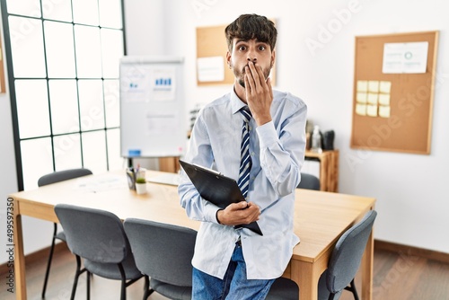 Hispanic man with beard wearing business style sitting on desk at office covering mouth with hand, shocked and afraid for mistake. surprised expression
