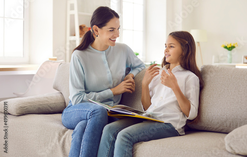 Happy mother and child talking while reading book together. Mom and daughter sitting on sofa at home and discussing interesting story. Au pair girl and kid practicing conversation in foreign language