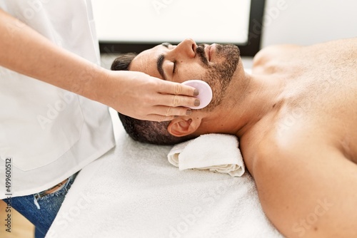 Young hispanic man having clean facial treatment using sponge at beauty center