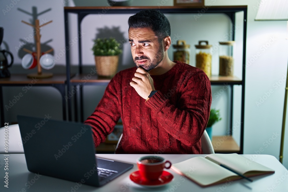 Young hispanic man with beard using computer laptop at night at home thinking worried about a question, concerned and nervous with hand on chin
