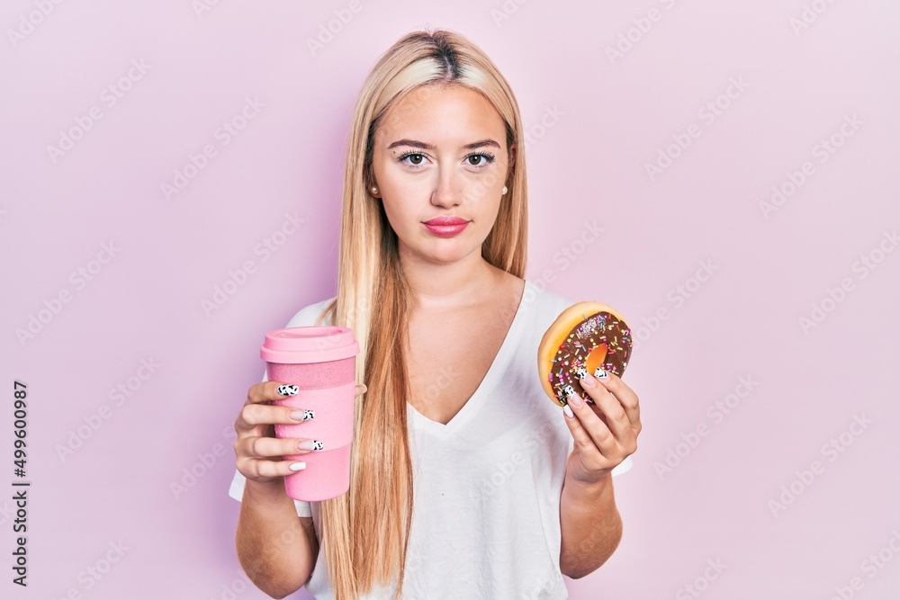 Young blonde girl eating doughnut and drinking coffee relaxed with serious expression on face. simple and natural looking at the camera. Young blonde girl eating doughnut and drinking coffee relaxed with serious expression on face. simple and natural looking at the camera.