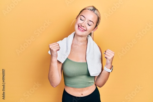 Young blonde girl wearing sportswear and towel very happy and excited doing winner gesture with arms raised, smiling and screaming for success. celebration concept.
