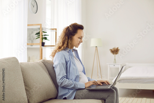 Young pregnant woman working, searching for information or shopping online using laptop. Side view of caucasian woman in third trimester of pregnancy sitting with laptop on sofa in living room at home