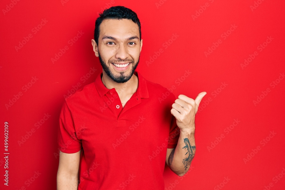 Fototapeta premium Hispanic man with beard wearing casual red t shirt smiling with happy face looking and pointing to the side with thumb up.