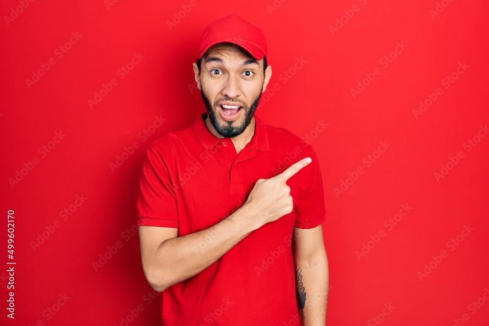 Fototapeta premium Hispanic man with beard wearing delivery uniform and cap surprised pointing with finger to the side, open mouth amazed expression.