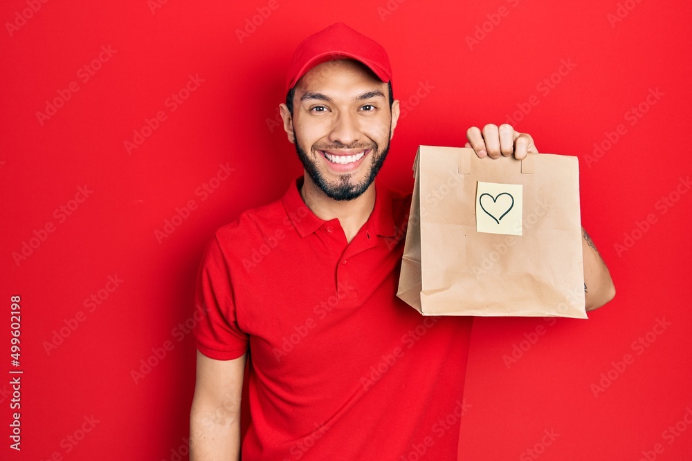Hispanic man with beard holding take away paper bag with heart reminder looking positive and happy standing and smiling with a confident smile showing teeth Hispanic man with beard holding take away paper bag with heart reminder looking positive and happy standing and smiling with a confident smile showing teeth