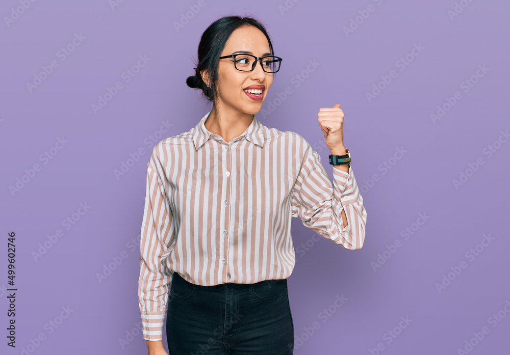 Young hispanic girl wearing casual clothes and glasses smiling with happy face looking and pointing to the side with thumb up. Young hispanic girl wearing casual clothes and glasses smiling with happy face looking and pointing to the side with thumb up.