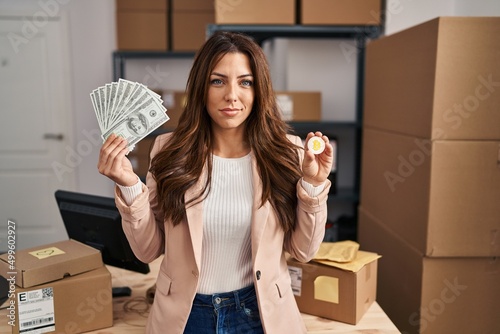 Fotografija Young brunette woman working at small business ecommerce holding bitcoin and money relaxed with serious expression on face