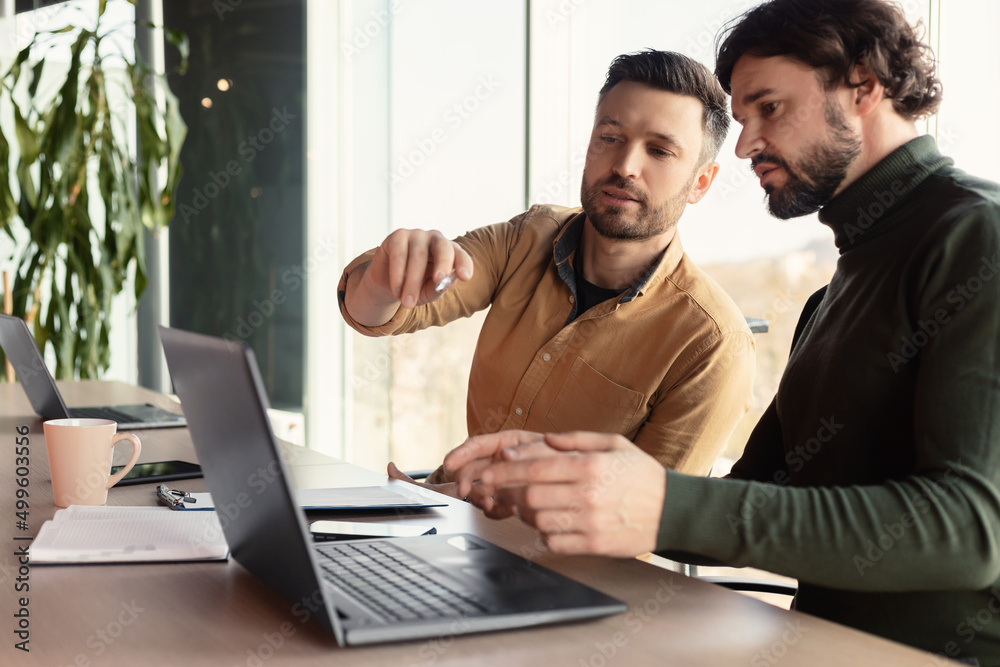 Obraz premium Serious employee speaking to his male colleague at company office, using laptop pc, discussing common business project