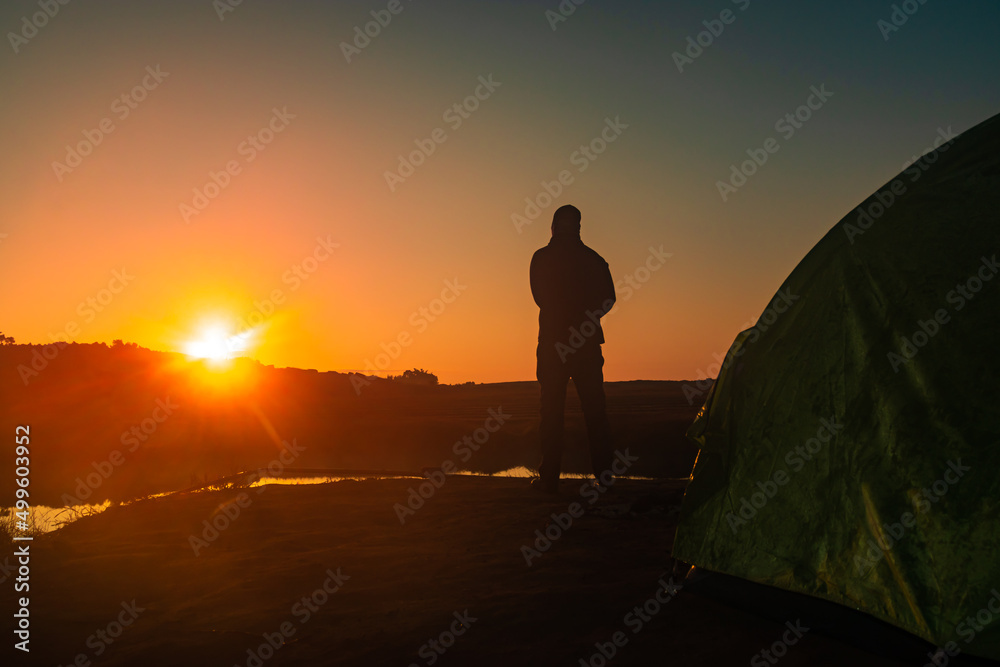 back lit man standing with sunrise over mountain shadow and calm lake with reflection at morning