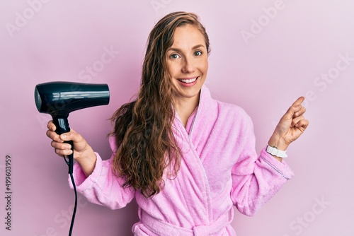 Young blonde woman wearing bathrobe using hair dryer smiling happy pointing with hand and finger to the side