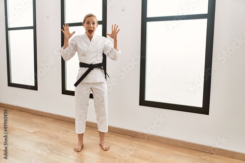 Caucasian young blonde woman wearing karate kimono and black belt celebrating victory with happy smile and winner expression with raised hands