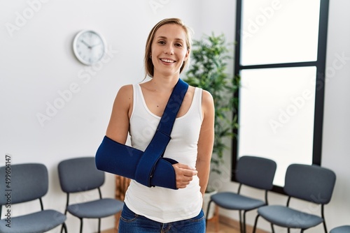 Young caucasian woman wearing sling on arm for accident standing at clinic waiting room photo