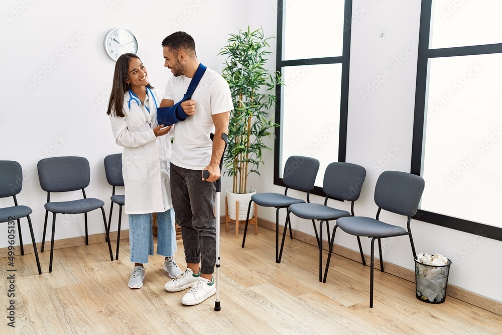 Youg latin injuried man wearing sling and using crutches talking with the doctor at clinic waiting room. Youg latin injuried man wearing sling and using crutches talking with the doctor at clinic waiting room.